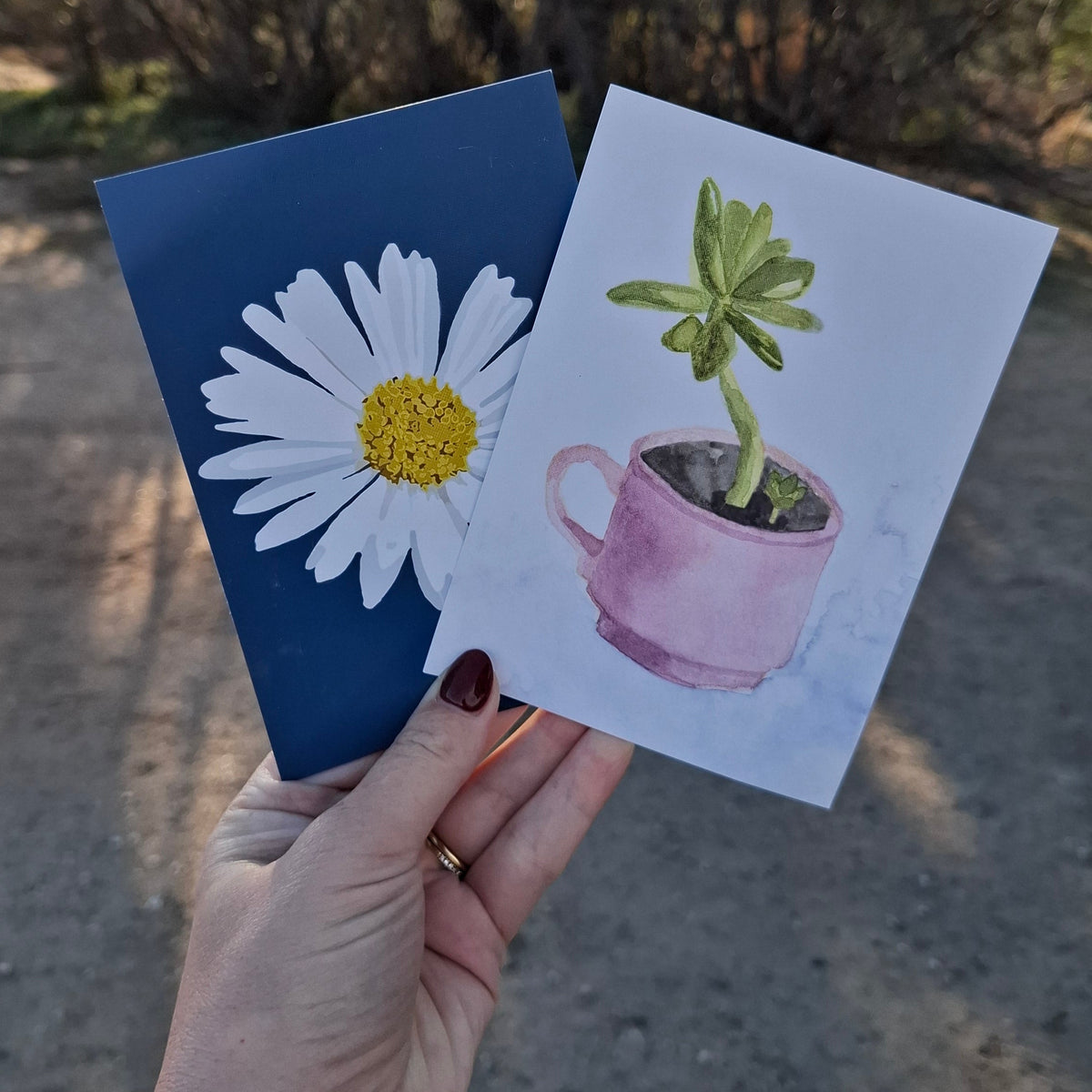Hand holding two small cards with plant illustrations against a natural background